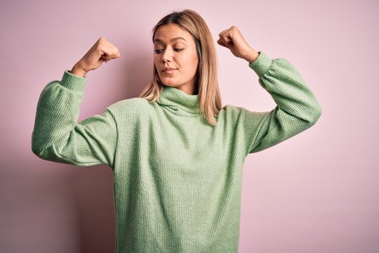 Young beautiful blonde woman wearing winter wool sweater over pink isolated background showing arms muscles smiling proud. Fitness concept.