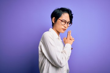 Young beautiful asian girl wearing casual shirt and glasses standing over purple background Holding symbolic gun with hand gesture, playing killing shooting weapons, angry face