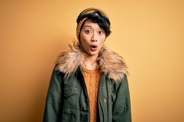 Beautiful asian skier girl wearing snow sportswear using ski goggles over yellow background afraid and shocked with surprise expression, fear and excited face.
