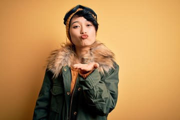 Beautiful asian skier girl wearing snow sportswear using ski goggles over yellow background looking at the camera blowing a kiss with hand on air being lovely and sexy. Love expression.