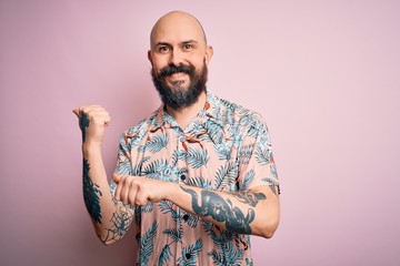 Handsome bald man with beard and tattoo wearing casual floral shirt over pink background Pointing...