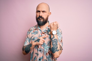 Handsome bald man with beard and tattoo wearing casual floral shirt over pink background In hurry pointing to watch time, impatience, looking at the camera with relaxed expression