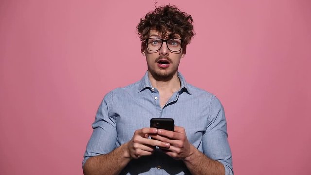 young casual man wearing glasses standing and reading something on phone surprised and then worrying and making a crazy sign pointing to the phone smiling in the end on pink studio background