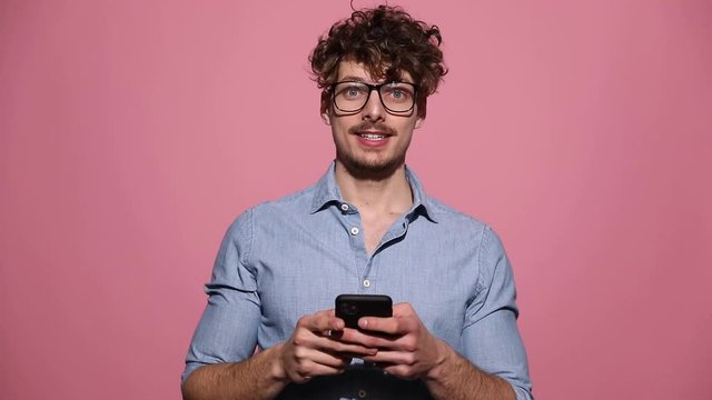 attractive casual man wearing glasses standing and texting on phone surprised then celebrating succes on pink studio background
