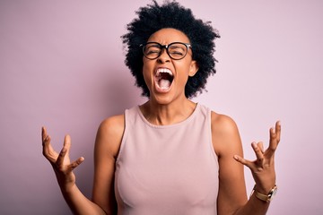 Young beautiful African American afro woman with curly hair wearing t-shirt and glasses crazy and mad shouting and yelling with aggressive expression and arms raised. Frustration concept.