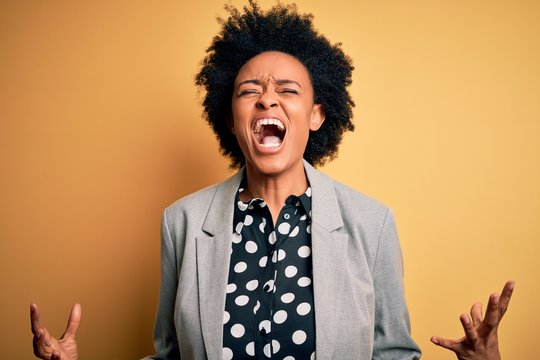 Young beautiful African American afro businesswoman with curly hair wearing jacket crazy and mad shouting and yelling with aggressive expression and arms raised. Frustration concept.