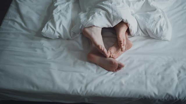 Bare feet of a loving couple lyuing under blanket and petting in bed. Intimate momennt of family life. Sex, relations concept. Filmed on RED camera.