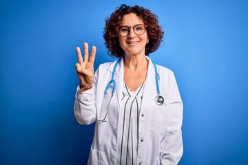 Middle age curly hair doctor woman wearing coat and stethoscope over blue background showing and pointing up with fingers number three while smiling confident and happy.