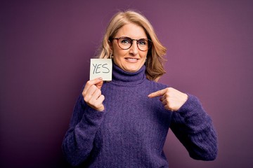 Middle age beautiful blonde woman wearing glasses holding reminder paper with yes message with...
