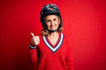Middle age beautiful blonde motorcyclist woman wearing moto helmet over red background doing happy thumbs up gesture with hand. Approving expression looking at the camera showing success.