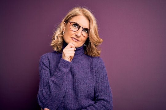 Middle age beautiful blonde woman wearing casual purple turtleneck sweater and glasses with hand on chin thinking about question, pensive expression. Smiling with thoughtful face. Doubt concept.