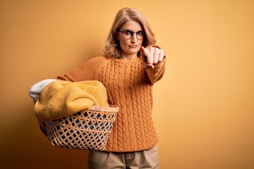 Middle age beautiful blonde woman doing housework holding wicker basket with clothes pointing with finger to the camera and to you, hand sign, positive and confident gesture from the front