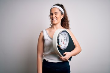 Young beautiful woman with curly hair holding weighing machine over white background winking...