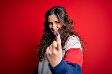 Fototapeta premium Young beautiful woman with curly hair wearing casual sweatshirt over isolated red background Beckoning come here gesture with hand inviting welcoming happy and smiling