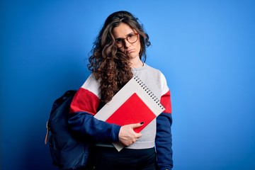Young beautiful student woman with curly hair wearing backpack holding book and notebook with a confident expression on smart face thinking serious