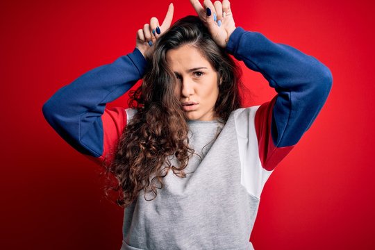 Young beautiful woman with curly hair wearing casual sweatshirt over isolated red background doing funny gesture with finger over head as bull horns