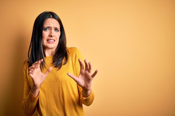 Young brunette woman with blue eyes wearing casual sweater over yellow background disgusted expression, displeased and fearful doing disgust face because aversion reaction. With hands raised
