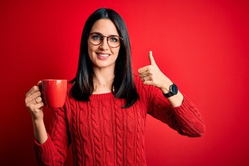 Young brunette woman with blue eyes wearing glasses and drinking a cup of coffee happy with big smile doing ok sign, thumb up with fingers, excellent sign