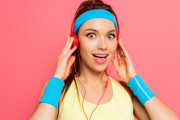 shocked sportswoman touching headphones while looking at camera isolated on pink