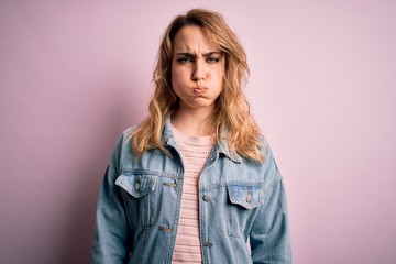 Young beautiful blonde woman wearing casual denim jacket standing over pink background puffing cheeks with funny face. Mouth inflated with air, crazy expression.