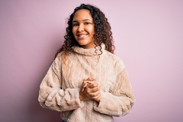 Young beautiful woman with curly hair wearing casual sweater standing over pink background with hands together and crossed fingers smiling relaxed and cheerful. Success and optimistic