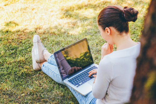 Mobile Office. Freelance business concept. Young woman sitting on green grass lawn in city park working on laptop pc computer. Lifestyle authentic candid student girl studying outdoors.