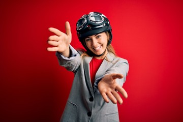 Young beautiful blonde motorcyclist woman wearing motorcycle helmet over red background looking at the camera smiling with open arms for hug. Cheerful expression embracing happiness.