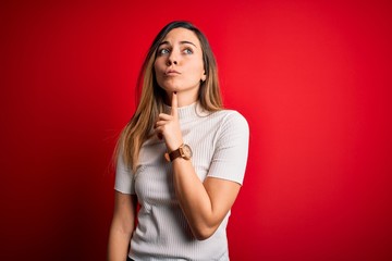 Beautiful blonde woman with blue eyes wearing casual white t-shirt over red background Thinking...