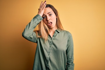 Young beautiful blonde woman with blue eyes wearing green shirt over yellow background surprised with hand on head for mistake, remember error. Forgot, bad memory concept.
