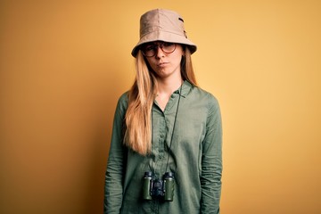 Beautiful blonde explorer woman with blue eyes wearing hat and glasses using binoculars skeptic and...