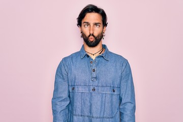 Young handsome hispanic bohemian man wearing hippie style over pink background making fish face with lips, crazy and comical gesture. Funny expression.