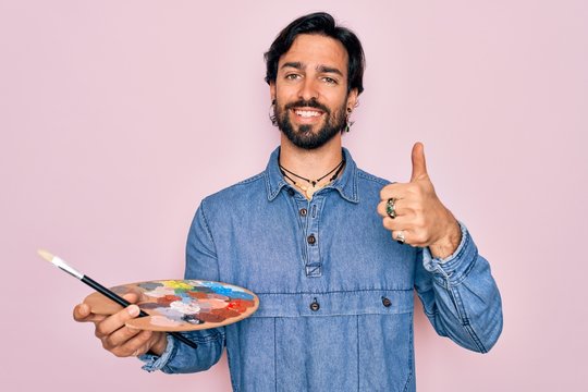 Young handsome hispanic artist man with bear holding painter palette and paintbrush happy with big smile doing ok sign, thumb up with fingers, excellent sign