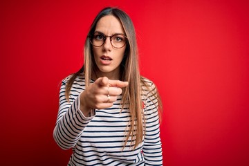 Young beautiful blonde woman with blue eyes wearing glasses standing over red background pointing displeased and frustrated to the camera, angry and furious with you