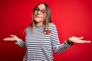 Young beautiful blonde woman with blue eyes wearing glasses standing over red background clueless...