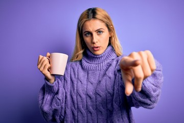 Young beautiful blonde woman drinking cup of coffee over isolated purple background pointing with finger to the camera and to you, hand sign, positive and confident gesture from the front