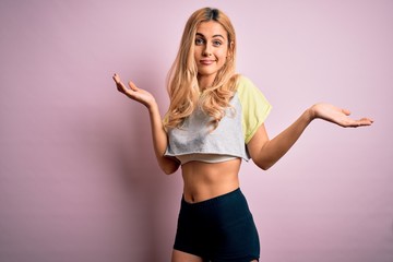Young beautiful blonde sporty woman doing sport wearing sportswear over pink background clueless and confused expression with arms and hands raised. Doubt concept.