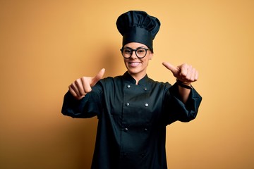 Young beautiful brunette chef woman wearing cooker uniform and hat over yellow background approving doing positive gesture with hand, thumbs up smiling and happy for success. Winner gesture.