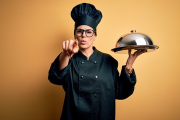 Young beautiful brunette chefwoman wearing cooker uniform and hat holding tray with dome pointing with finger to the camera and to you, hand sign, positive and confident gesture from the front