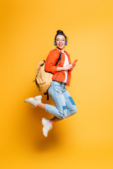 excited student in headset and glasses jumping while holding smartphone on yellow background - obrazy, fototapety, plakaty
