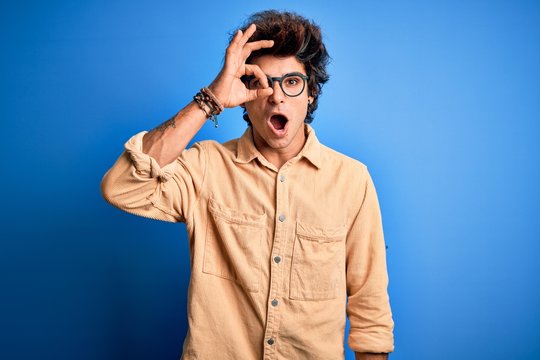 Young handsome man wearing casual shirt standing over isolated blue background doing ok gesture shocked with surprised face, eye looking through fingers. Unbelieving expression.