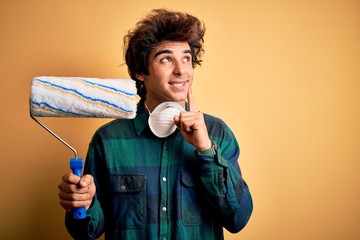 Young handsome painter man holding roller standing over isolated yellow background serious face...