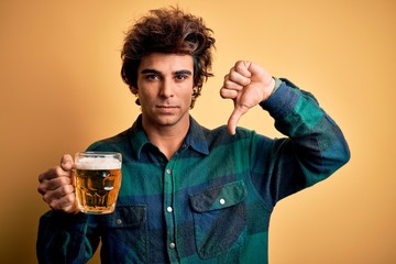 Young handsome man drinking jar of beer standing over isolated yellow background with angry face,...