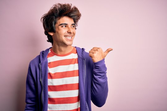 Young man wearing striped t-shirt and purple sweashirt over isolated pink background smiling with happy face looking and pointing to the side with thumb up.