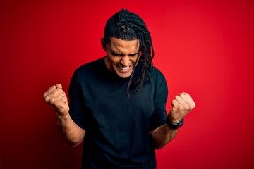 Fototapeta premium Young handsome african american afro man with dreadlocks wearing casual t-shirt very happy and excited doing winner gesture with arms raised, smiling and screaming for success. Celebration concept.