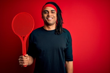 African american afro sportsman with dreadlocks holding tennis racket over red background with a happy face standing and smiling with a confident smile showing teeth