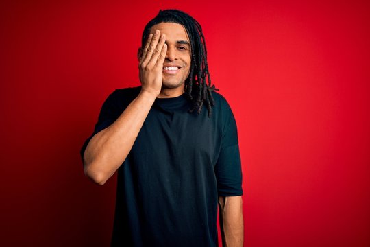 Young handsome african american afro man with dreadlocks wearing casual t-shirt covering one eye with hand, confident smile on face and surprise emotion.