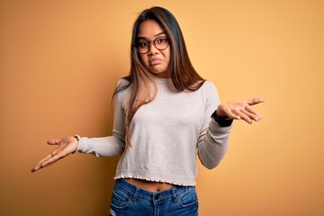 Young beautiful asian girl wearing casual sweater and glasses over yellow background clueless and...
