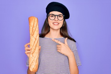 Young beautiful girl wearing fashion french beret holding fresh baked bread baguette very happy pointing with hand and finger