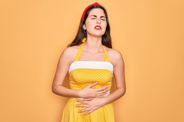 Young beautiful pin up woman wearing 50s fashion vintage dress over yellow background with hand on...