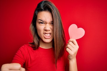 Young beautiful brunette romantic girl holding red paper heart shape over isolated background annoyed and frustrated shouting with anger, crazy and yelling with raised hand, anger concept